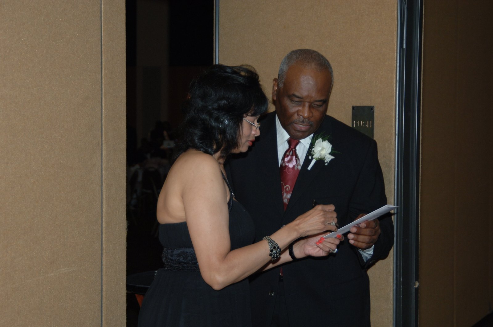 Two people at formal event - woman in black dress on left, man in black suit with red tie and white flower on right, both looking at document